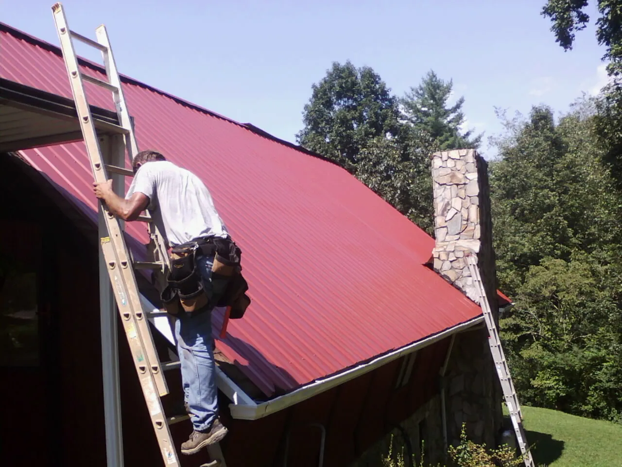 Roofer on a ladder beside a red metal roof and stone chimney — Soffit & Fascia Repair by Roofing Brothers in Reedsport